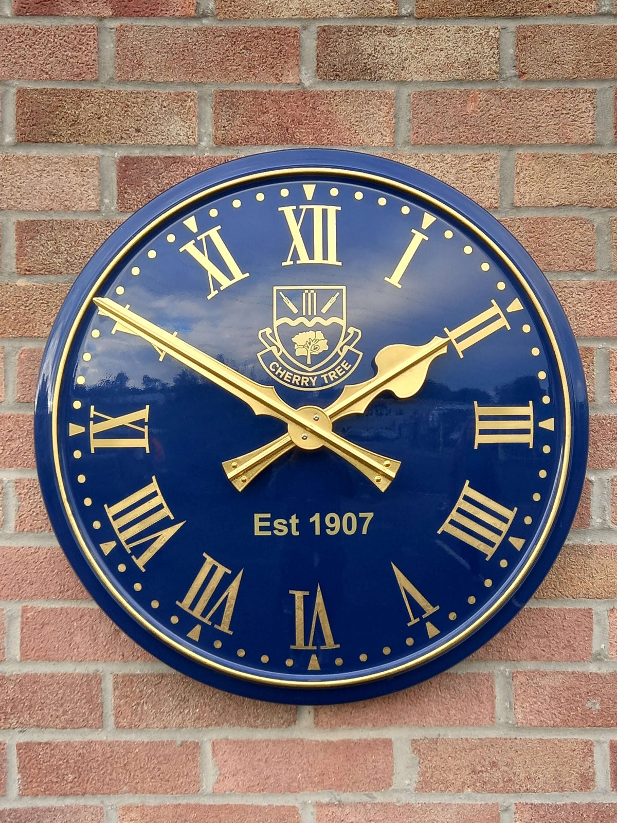 Cricket Pavilion Clocks with blue face, gold hands and numerals, mounted on a brick wall.