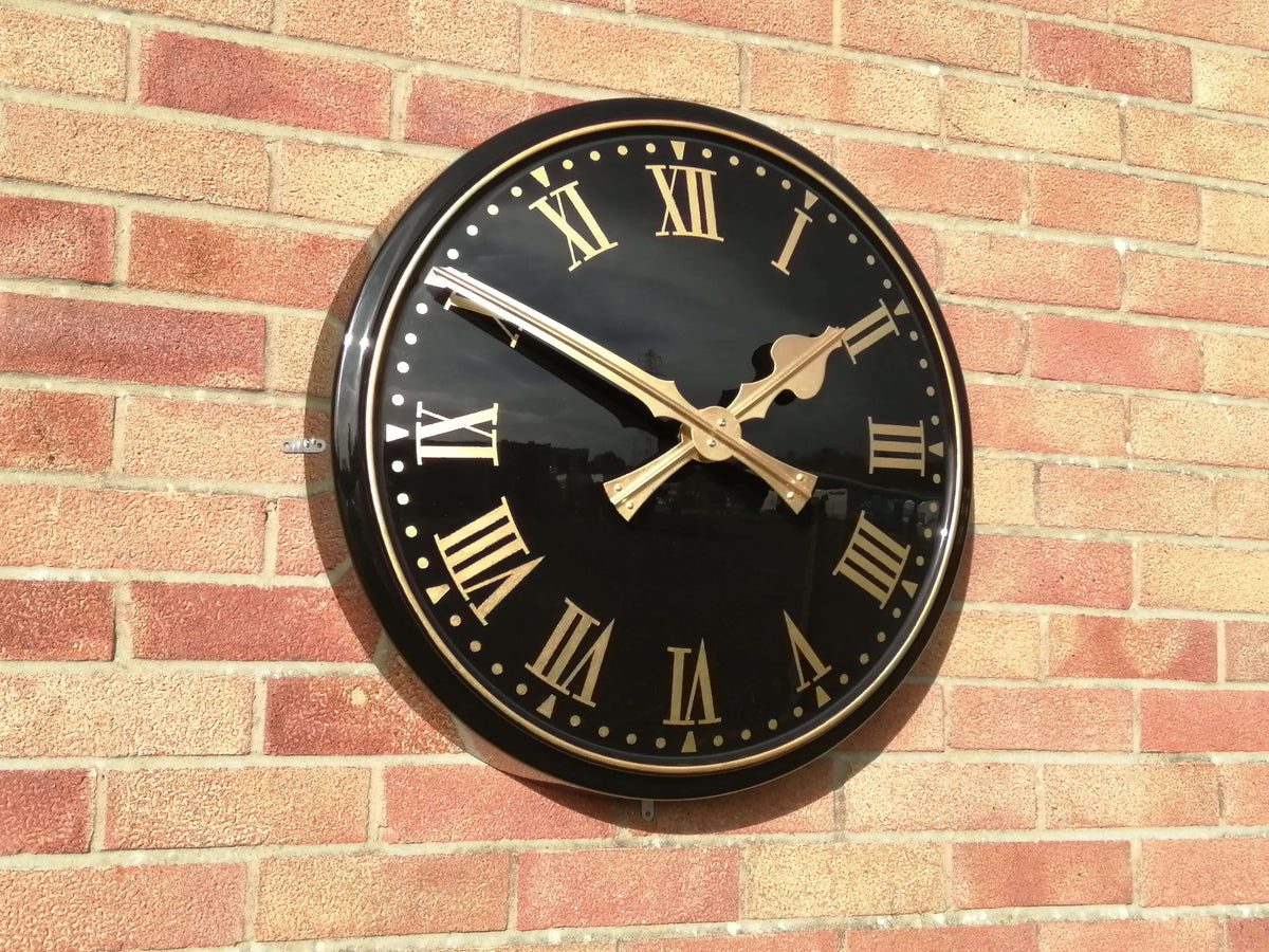 Cricket Pavilion Clocks traditional wall clock with black dial and gold hands mounted on a brick wall.