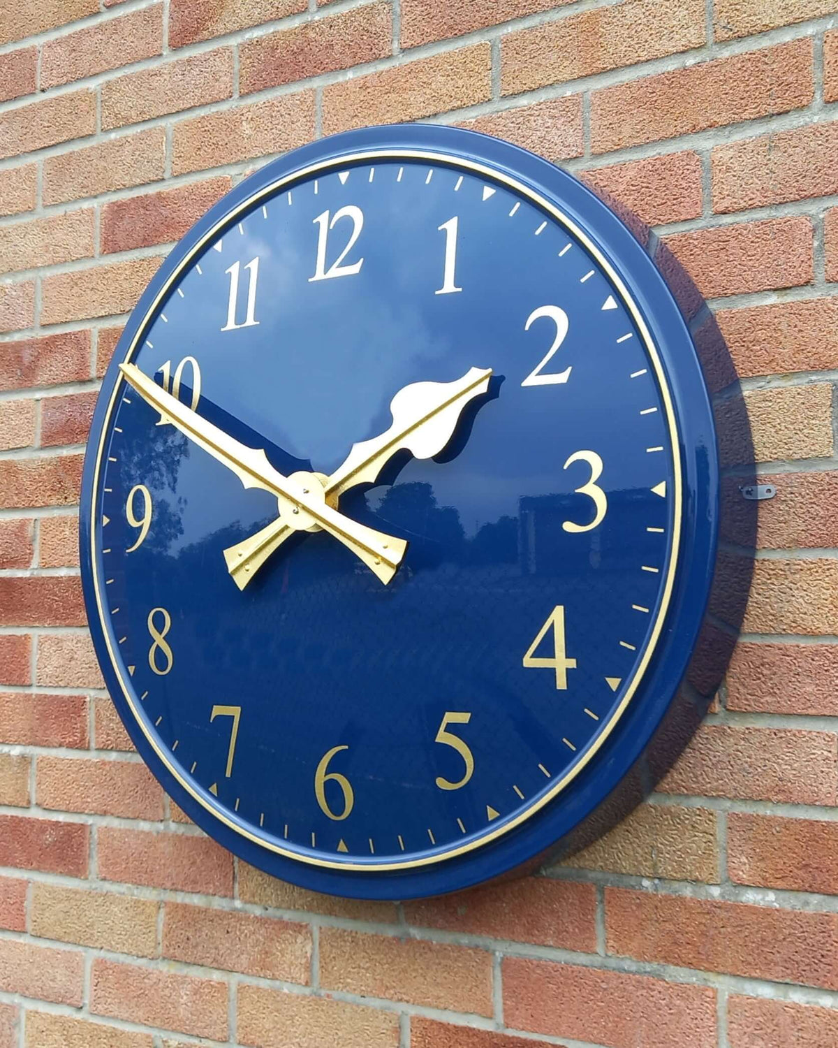 Cricket Pavilion Clocks - traditional wall clock with navy blue dial and gold hands mounted on a brick wall.